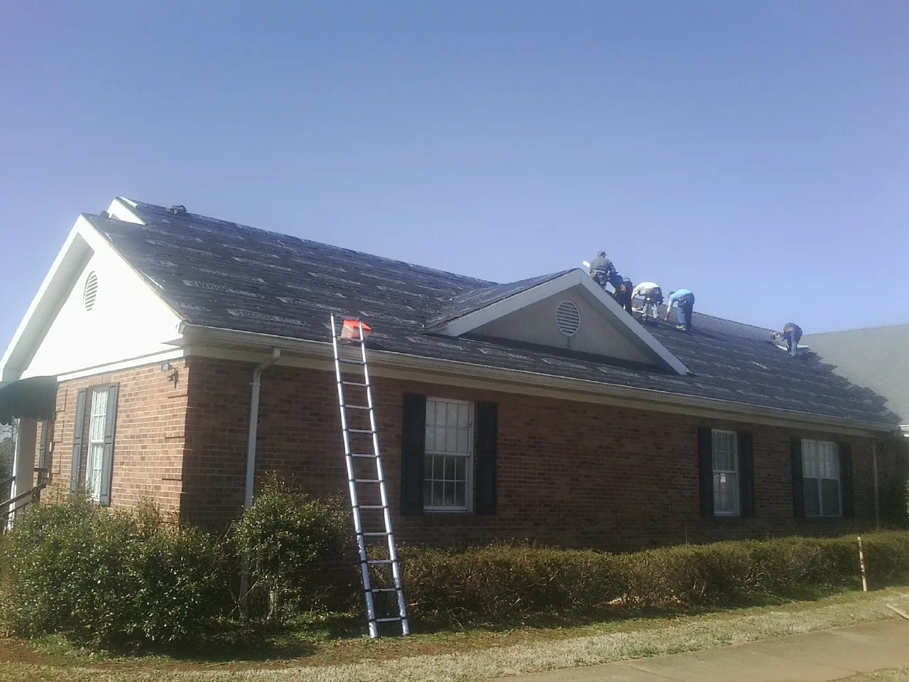 Crew tearing shingles off a brick ranch roof with a ladder set — Emergency Roof Repair work by our crew in East Thetford
